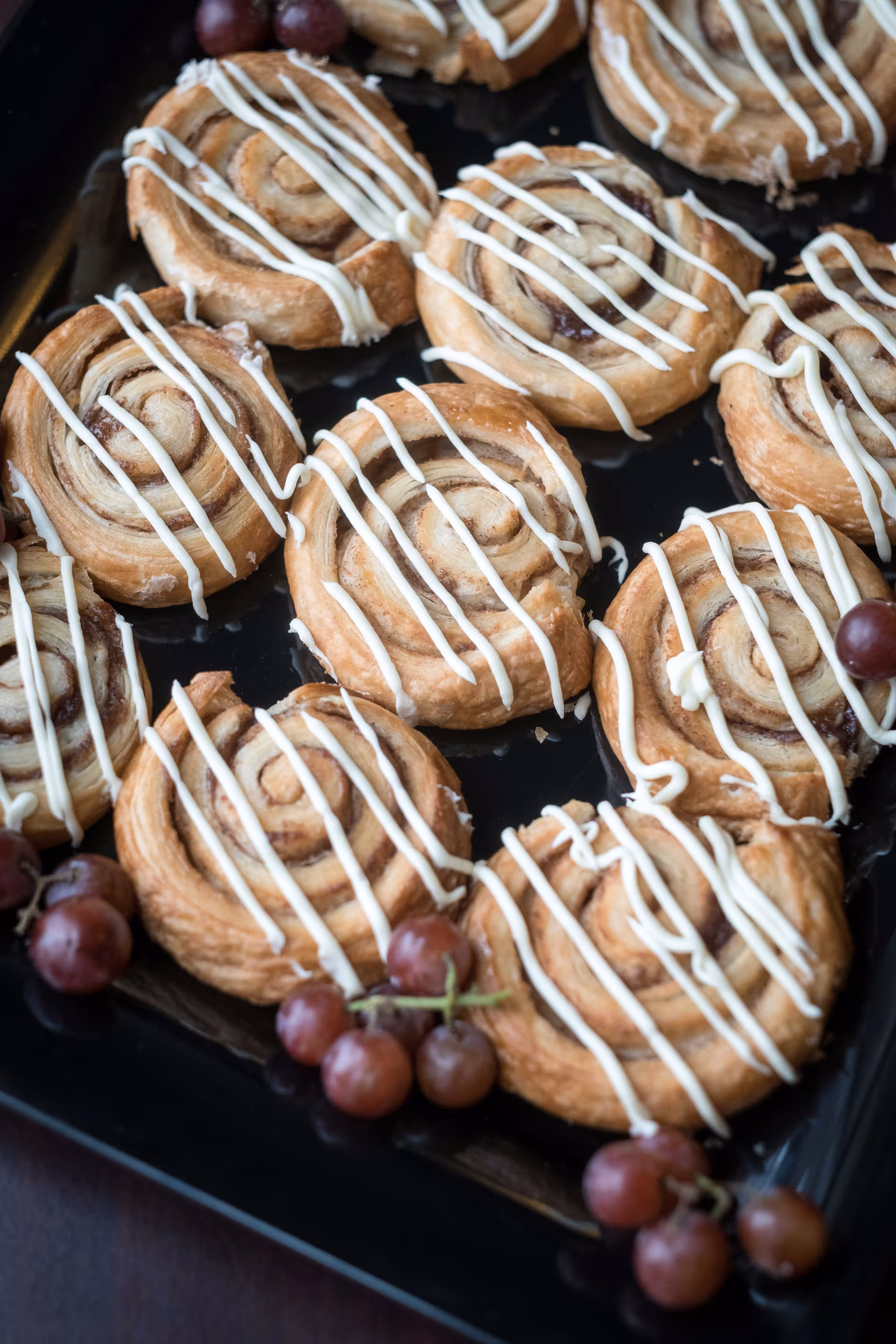 A close-up view of a tray filled with cinnamon rolls drizzled with white icing, garnished with small bunches of red grapes.