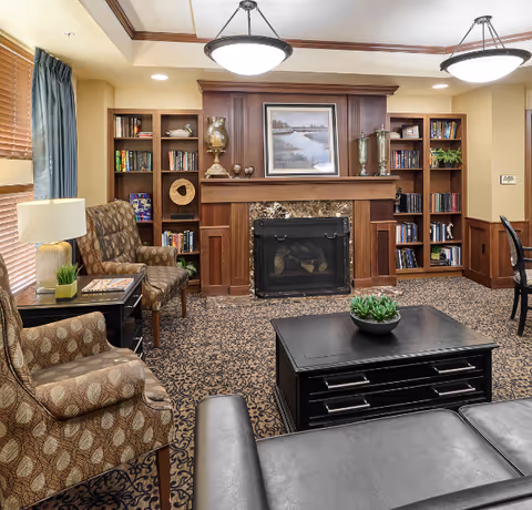 A cozy living room with patterned carpet, two upholstered armchairs, a black leather sofa, and a black coffee table with a small green plant. The room features a wooden fireplace with a marble surround, flanked by built-in bookshelves filled with books and decorative items. Two ceiling lights hang from the ceiling, and there is a window with wooden blinds and blue curtains on the left side.