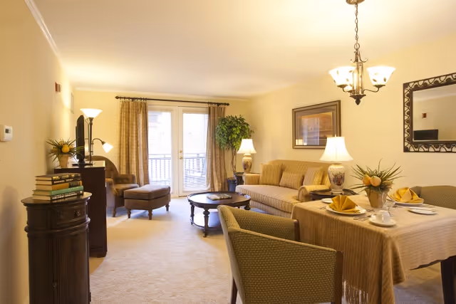 A cozy living room and dining area in a senior living community. The living room features a beige sofa, an armchair with an ottoman, a coffee table, a floor lamp, and a potted plant near glass doors with curtains. The dining area has a table covered with a beige tablecloth, set with plates, napkins, cups, and a floral centerpiece. The walls are light-colored with framed artwork and a decorative mirror.