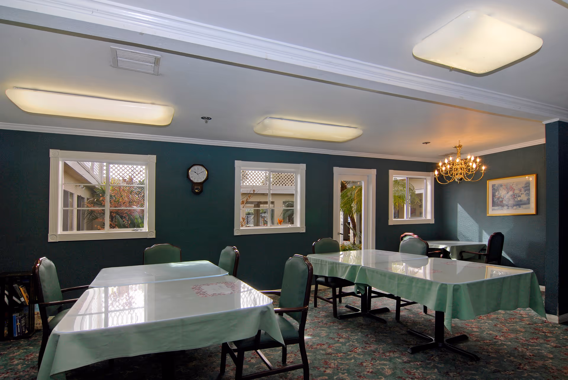 Interior view of a dining room with multiple tables covered in green tablecloths and chairs around them. The room has dark green walls, three windows, a wall clock, a chandelier, and a framed picture on the wall. There is a door leading outside with visible plants through the windows and door.