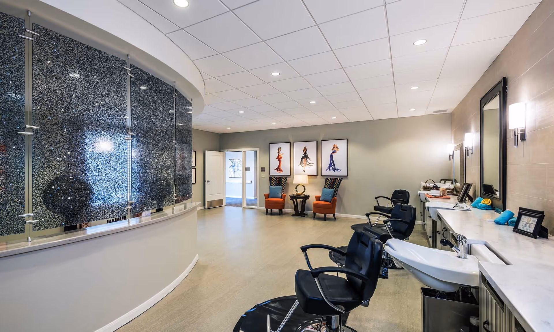 A modern salon area inside a senior living facility featuring three black salon chairs in front of a long counter with sinks and mirrors. The room has a curved wall with a textured glass panel, two orange armchairs with a small table between them, and three framed fashion illustrations on the far wall. The space is well-lit with recessed ceiling lights and wall sconces.