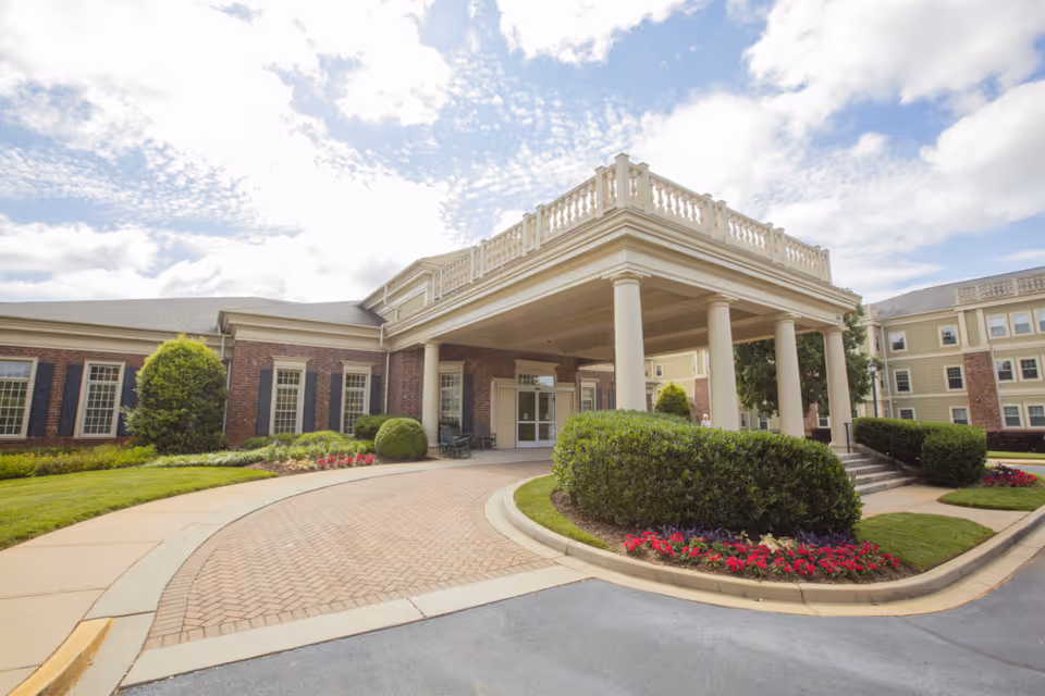 The exterior entrance of The Woodlands at Furman senior living facility featuring a covered drop-off area with large white columns, brick walls, manicured bushes, and flower beds under a partly cloudy sky.