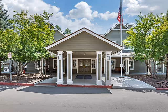 Front entrance of a senior living facility with a covered driveway, two white pillars, an American flag on a flagpole, and green trees on either side under a partly cloudy sky.