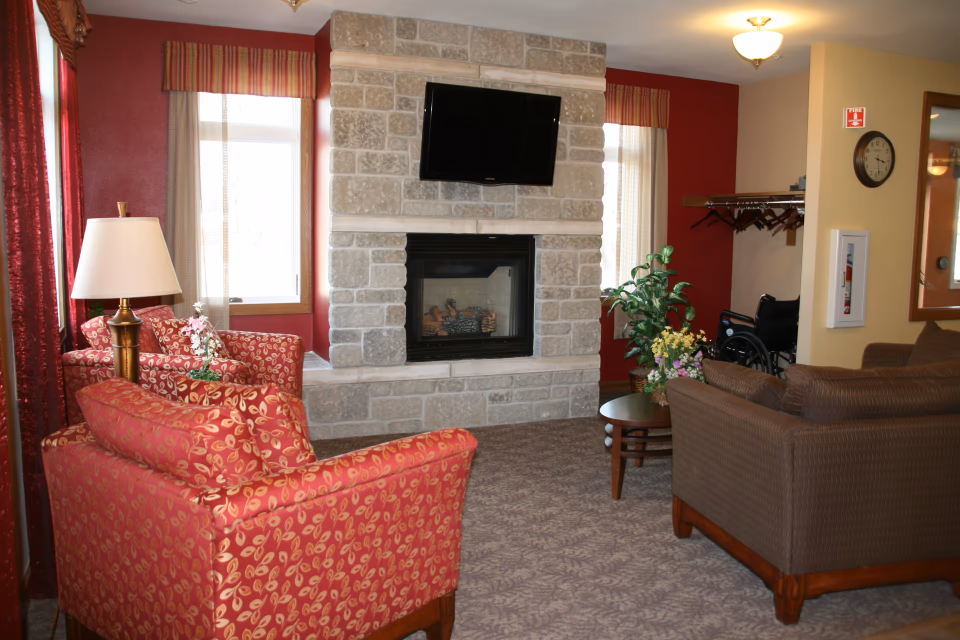A cozy living room area with a stone fireplace and a mounted flat-screen TV above it. The room features red patterned armchairs, a brown sofa, a wooden coffee table with a floral arrangement, a floor lamp, and windows with striped valances. A wheelchair is visible near a coat rack on the right side of the room.