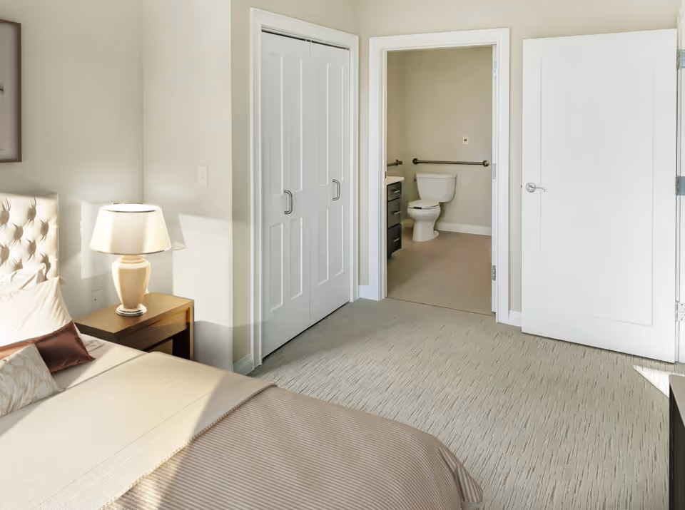 A bright bedroom with a bed, nightstand and lamp, double closet doors, and an open doorway showing a bathroom with a toilet and grab bar.