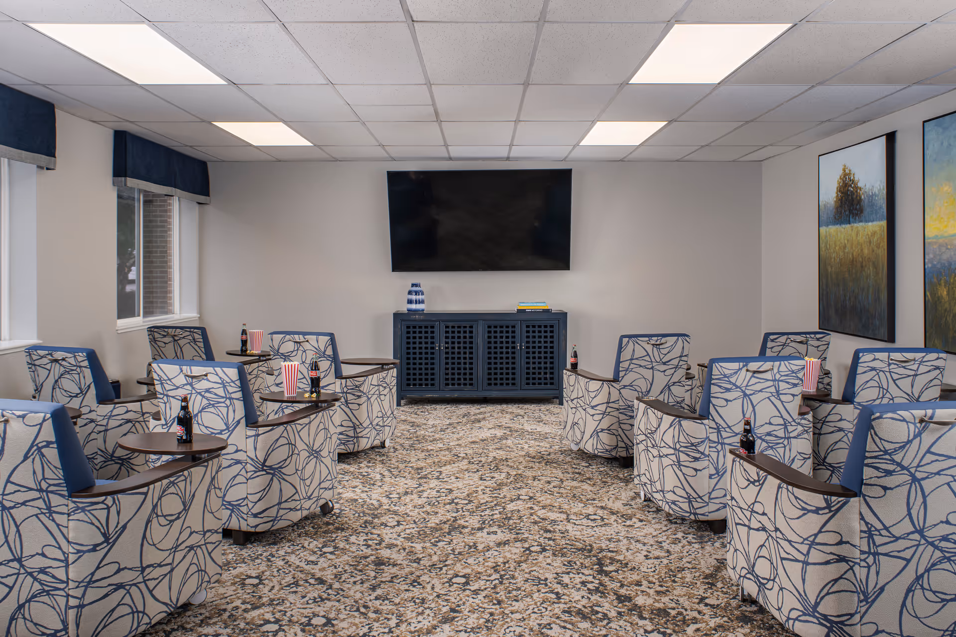 A cozy common room with patterned armchairs arranged in two rows facing a wall-mounted flat screen TV. Each chair has a small attached table holding a bottle of soda and a popcorn container. The room has a carpeted floor, two large landscape paintings on the right wall, and windows with blue valances on the left.