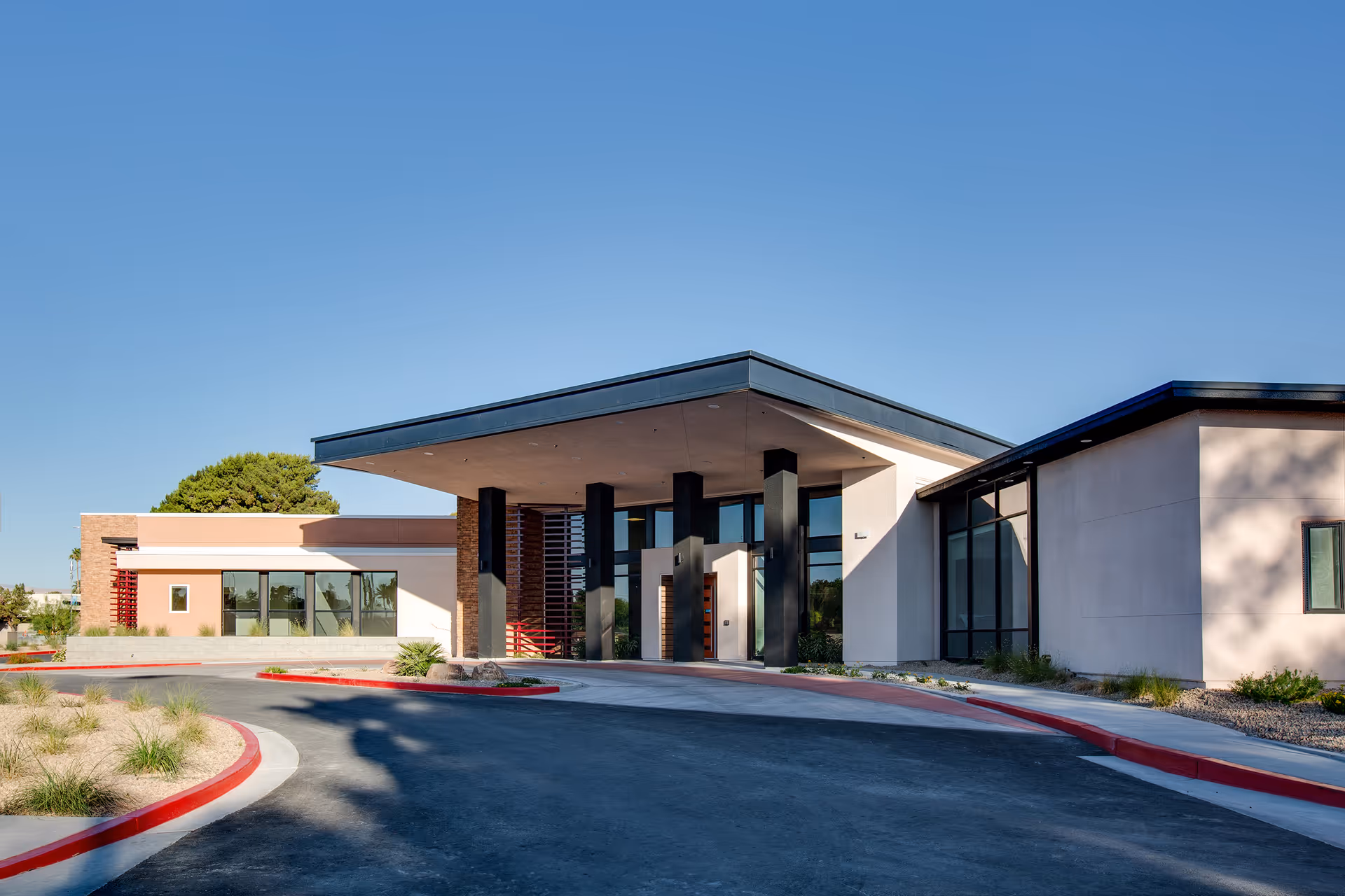 Modern single-story building entrance with a large covered porte-cochere and a curved driveway under a clear blue sky.