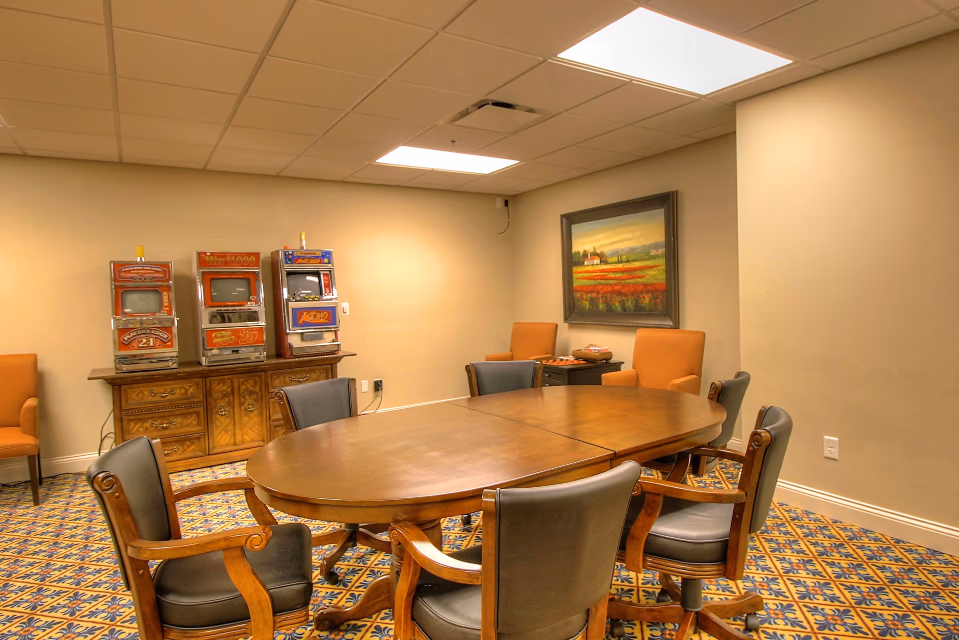 Conference-style room with an oval wooden table surrounded by chairs, a cabinet with slot machines, and a framed landscape painting on the wall.
