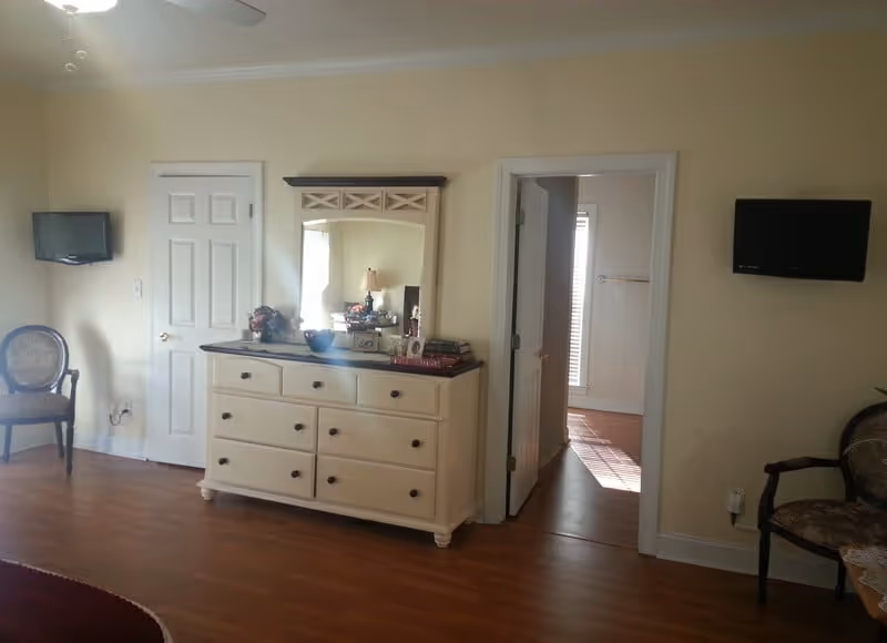 Interior of a senior living room with dresser, mirror, chairs, and wall-mounted TVs.