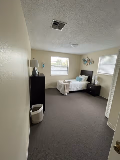 A small, simple bedroom with a single bed against the far wall, decorated with white bedding and blue accent pillows. There is a dark wooden nightstand with a lamp next to the bed, and a matching dresser with a lamp and decorative items on top. Two windows with blinds allow natural light into the room. The walls are light-colored, and the floor is carpeted in gray. A white basket is placed near the dresser.