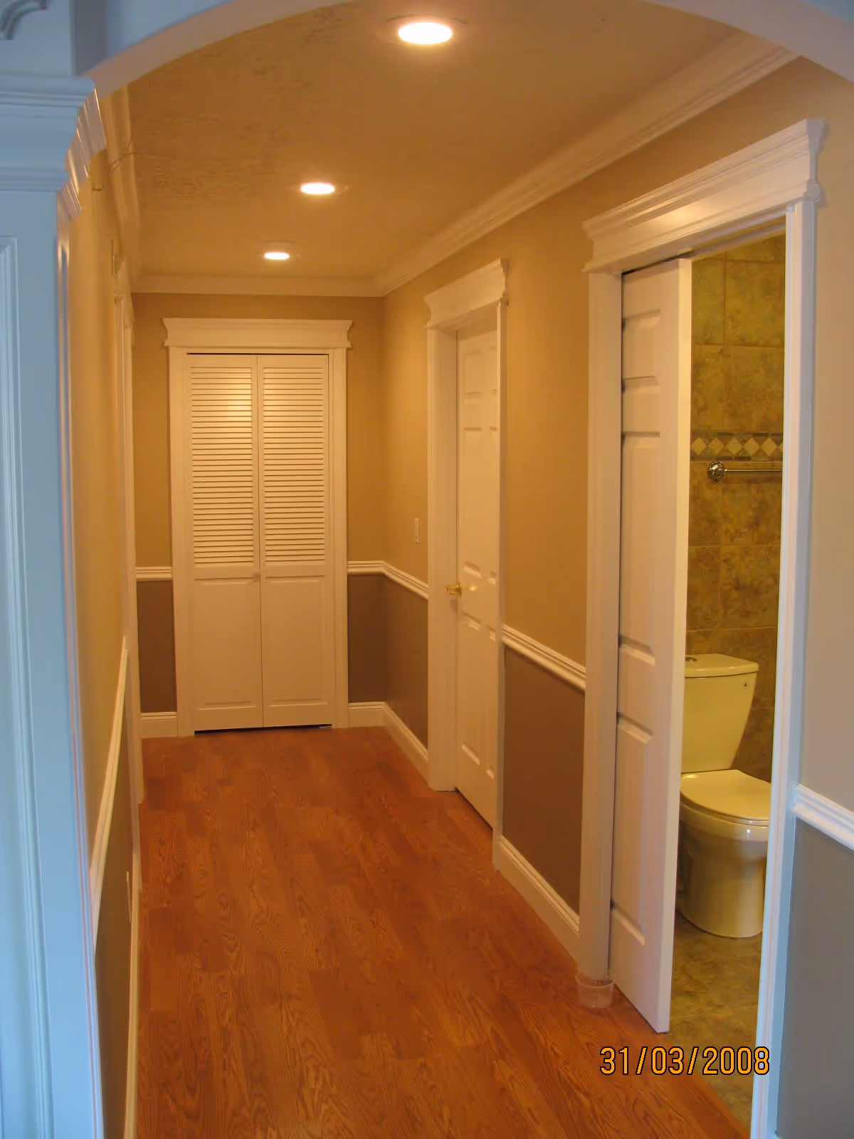 Well-lit interior hallway with wood floors, white doors, and a partially visible bathroom with a toilet.