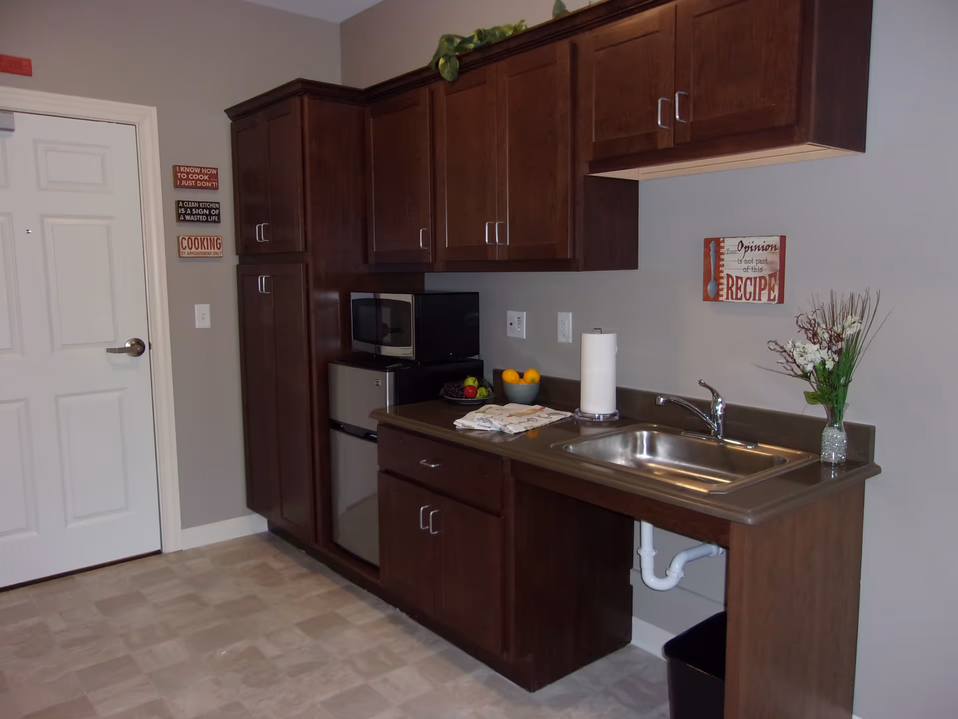 A small kitchen area with dark wood cabinets, a stainless steel sink, a microwave, a mini refrigerator, and a countertop with a bowl of fruit, paper towel holder, and a vase with flowers. There are three decorative signs on the walls with cooking-related quotes. The floor is tiled and a white door is visible on the left side.