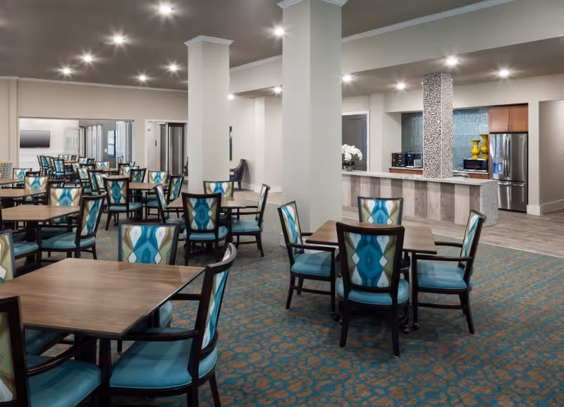 A spacious dining room with multiple wooden tables and chairs featuring blue and patterned upholstery. The room has a carpeted floor with a blue and brown pattern, white walls, and recessed ceiling lights. In the background, there is a kitchen area with stainless steel appliances, including a refrigerator and microwave, and a counter with decorative vases.