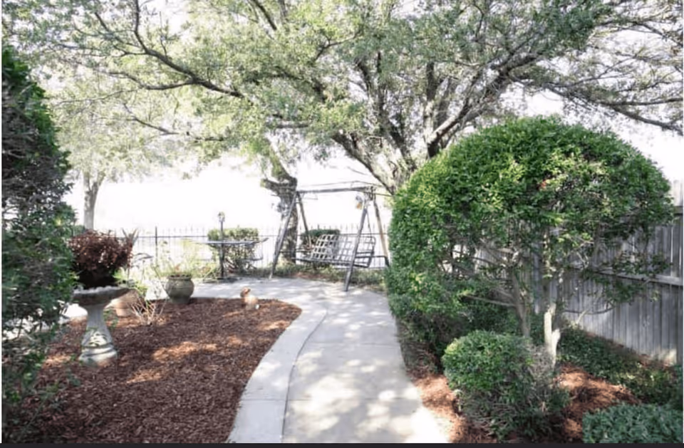 A peaceful outdoor garden area with a concrete pathway winding through mulch beds and trimmed bushes. There is a metal swing bench under a large tree providing shade, and a small round table with chairs in the background. The area is enclosed by a wooden fence.