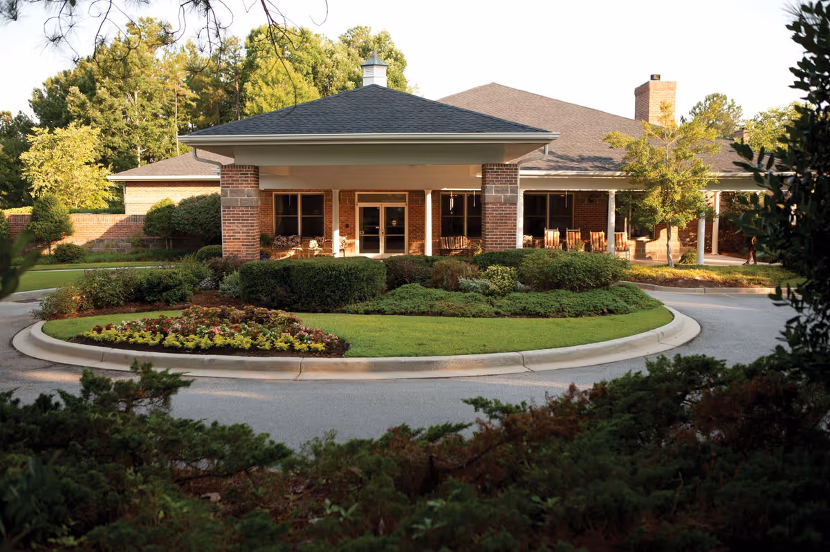 Front exterior view of a senior living facility with a covered entrance supported by brick columns, surrounded by well-maintained landscaping including bushes, flowers, and trees, with a circular driveway in front.