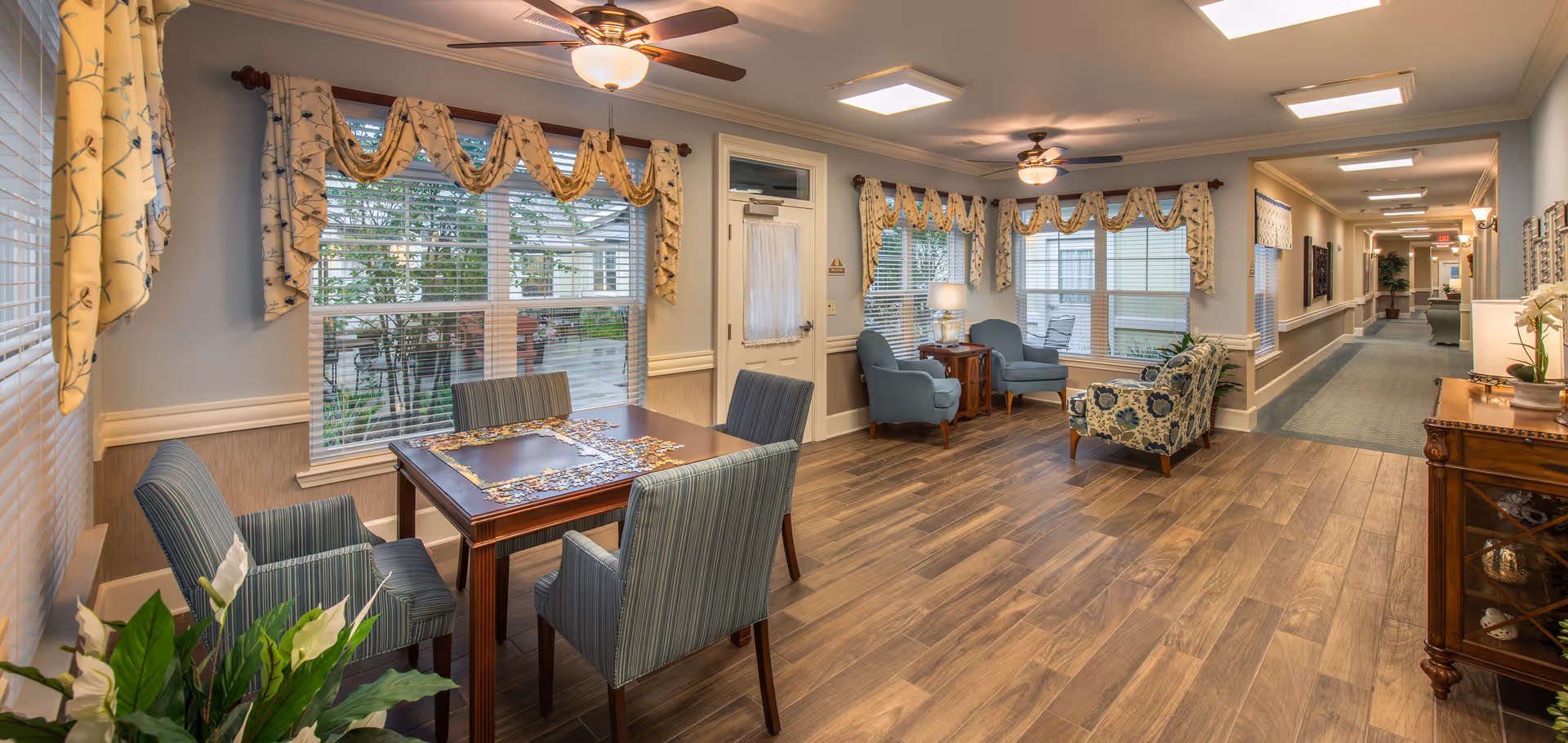 Well-lit senior living common area with a table and chairs, lounge seating, large windows, and a long hallway.