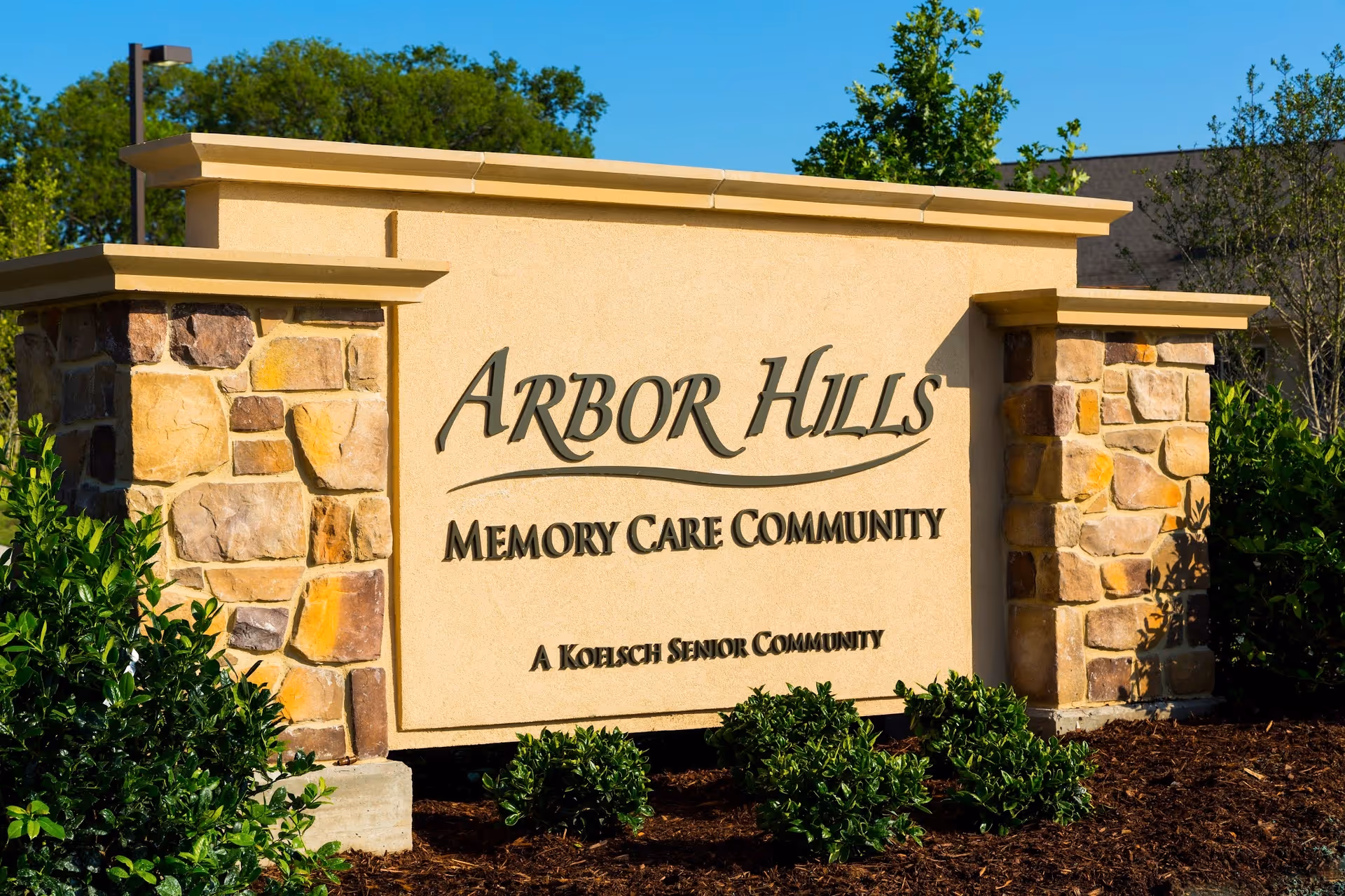 Stone and stucco sign for Arbor Hills Memory Care Community, a Koelsch Senior Community, surrounded by green shrubs and trees under a clear blue sky.