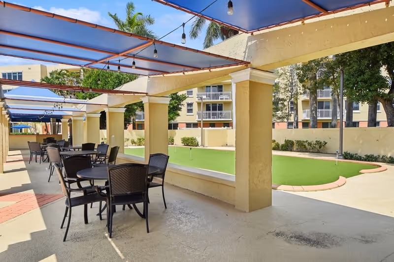 Outdoor covered patio area with multiple round tables and chairs, overlooking a small putting green. The patio has a blue canopy roof with string lights hanging underneath. In the background, there are palm trees and a multi-story residential building.