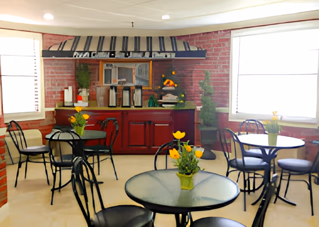 A cozy dining area with round glass tables and black metal chairs. Each table has a small green vase with yellow flowers. The room has red brick walls, two large windows with white blinds, and a red cabinet with a countertop against the back wall. On the countertop are coffee dispensers and a small decorative plant. Two tall green potted plants flank the cabinet.