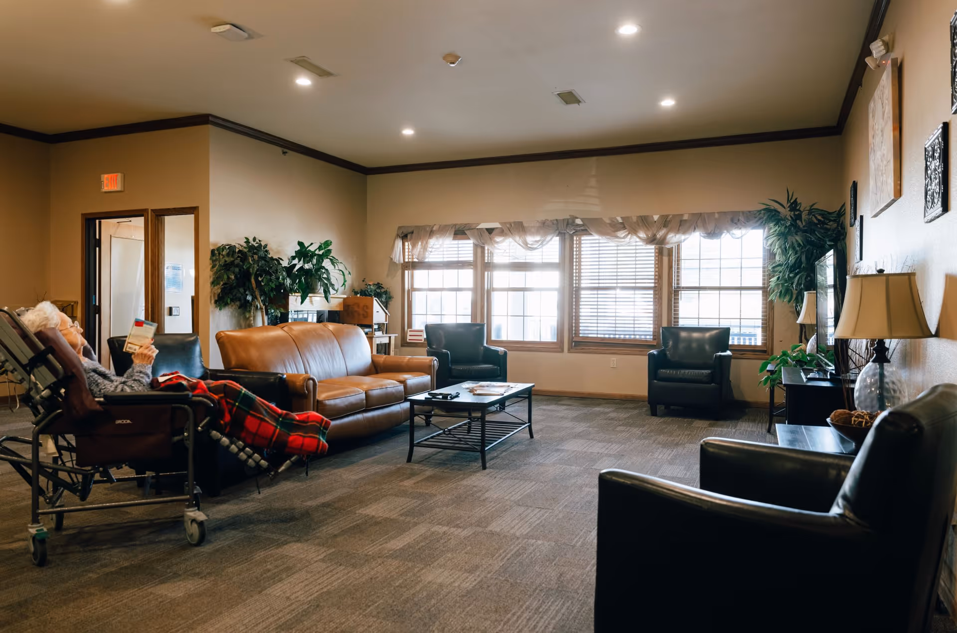 A spacious assisted-living common room with leather sofas and armchairs around a coffee table, large windows, plants, and a person reclining in a chair reading.