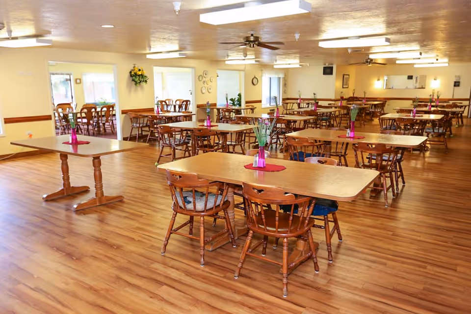 A spacious dining room with multiple wooden tables and chairs arranged neatly. Each table has a small pink vase with green plants on a red placemat. The room has wooden flooring, ceiling fans, and bright lighting. There are windows and doorways leading to other rooms in the background.