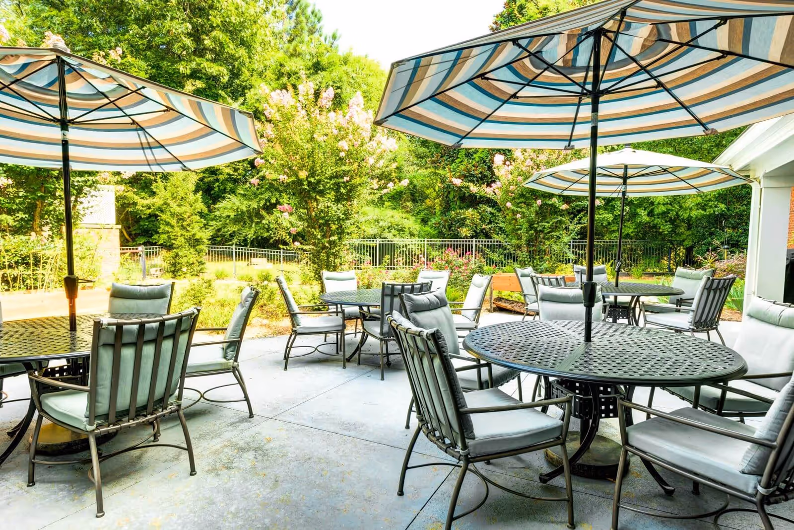 Outdoor patio area with several round metal tables and cushioned chairs under large striped umbrellas, surrounded by greenery and trees.