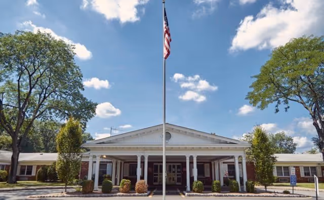 Front entrance of a single-story care facility with a flagpole, white columns, and landscaping under a blue sky.