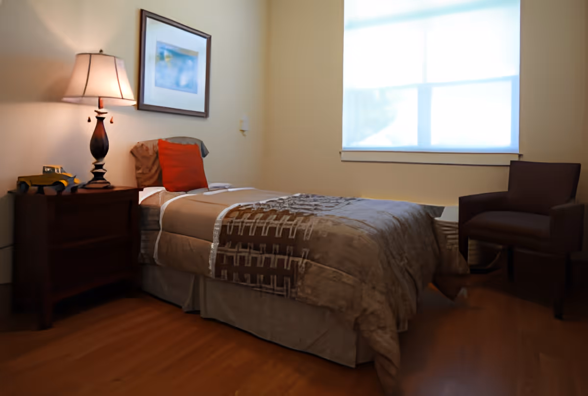 A cozy bedroom with a single bed covered in a brown and beige patterned comforter, a red pillow, a wooden nightstand with a lamp and a small decorative item, a framed picture on the wall, a window with a white blind, and a brown armchair.