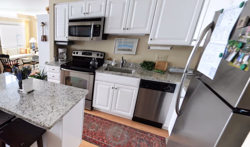 Bright kitchen with white cabinets, granite countertops, a small island, and stainless steel appliances opening to a living area.