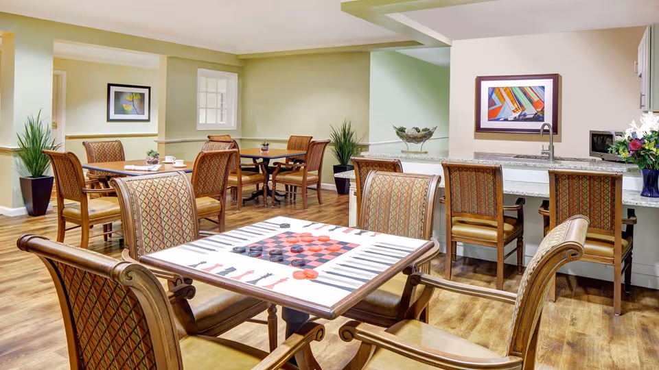 A well-lit common area with multiple tables and chairs, including a table in the foreground featuring a board game with checkers and chess pieces. The room has wooden flooring, light green and beige walls, framed artwork, and plants. There is a counter with bar stools and a sink in the background.