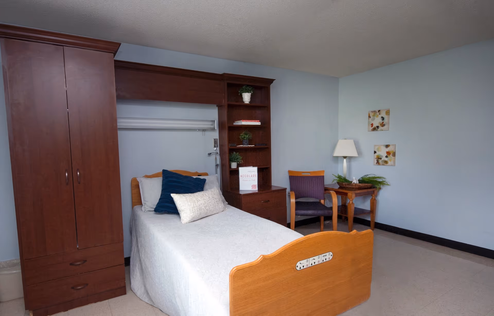 A single bed with a wooden headboard and footboard in a room with light blue walls. The bed is made with a white quilt and has three pillows, one dark blue and two light-colored. Next to the bed is a tall wooden wardrobe and a built-in wooden shelving unit with small plants and a welcome sign. There is a wooden chair with a purple cushion and a small wooden side table with a lamp and a plant. Two abstract paintings hang on the wall above the side table.