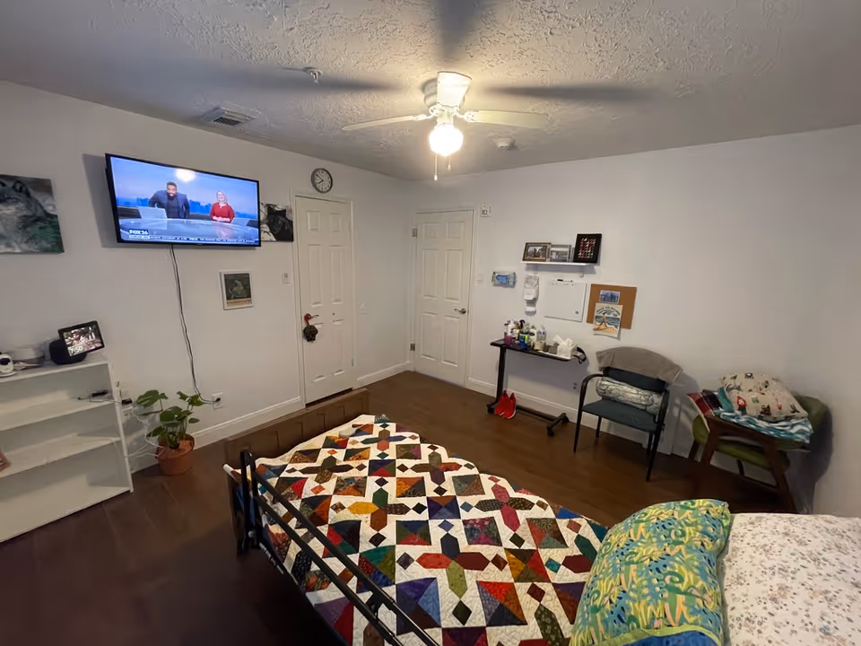 A senior living facility bedroom with a bed covered in a colorful patchwork quilt, a ceiling fan with a light, a wall-mounted TV showing a news program, two closed white doors, a small table with various items including hand sanitizer, a chair with folded blankets, and a small plant on the floor near a white shelf.