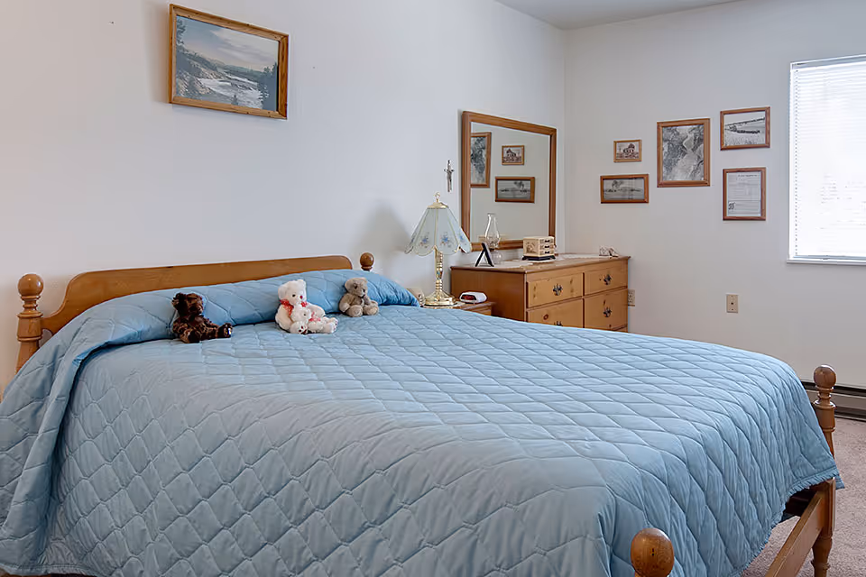 A bedroom with a wooden bed frame and a light blue quilted bedspread. Three teddy bears are placed on the bed. There is a wooden dresser with a mirror and a decorative lamp on top. Several framed pictures and documents hang on the white walls. A window with blinds allows natural light into the room.