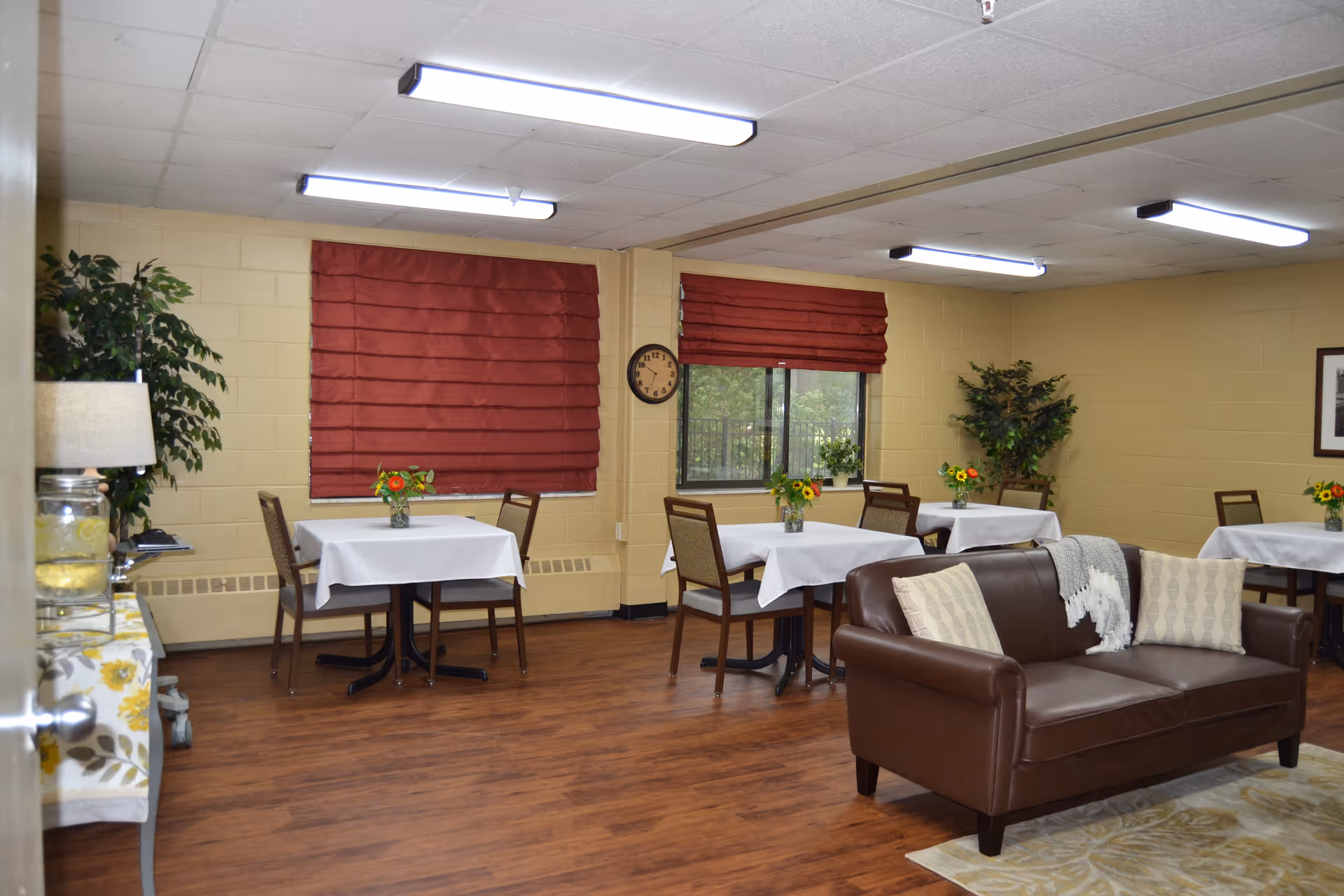 Communal dining and lounge area with small tables set with white tablecloths and vases, a brown leather sofa, and red window shades.