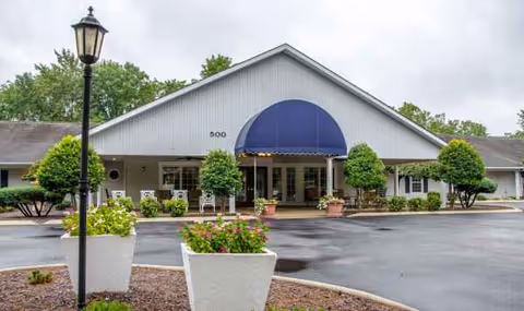 Single-story senior living facility with a blue awning over the entrance, surrounded by potted plants.