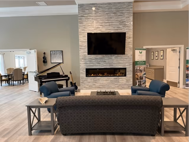 A lounge with a sofa and two armchairs facing a wall-mounted TV above a linear fireplace, with a piano and dining area visible to the left.