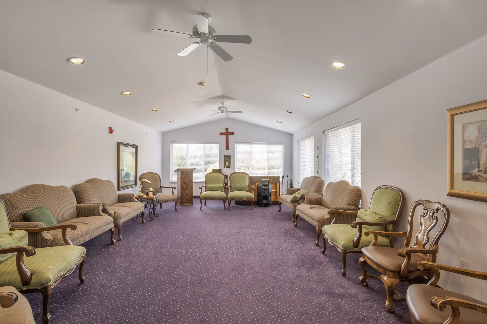 Carpeted communal sitting room with upholstered chairs arranged along the walls, a small wooden podium, and a cross on the far wall.