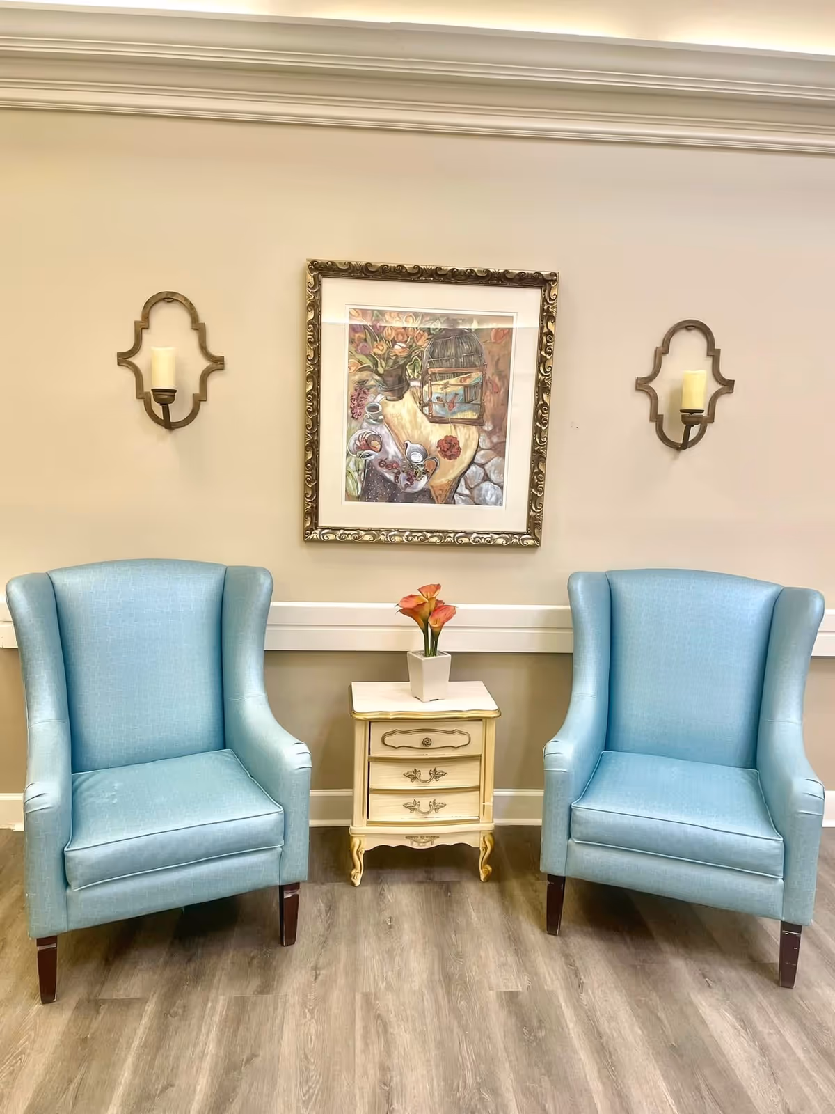 Two light-blue wingback chairs flank a small cream side table with a vase of flowers beneath framed artwork and wall sconces in a seating area.