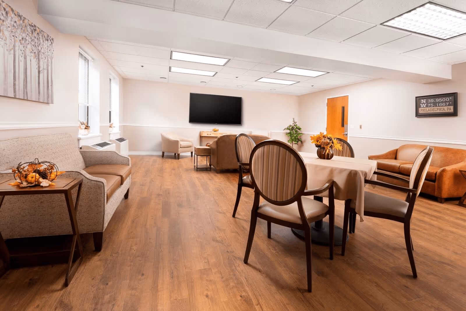 A spacious and well-lit common area with wooden flooring, featuring a round table with four chairs covered in beige upholstery, two sofas, an armchair, a wall-mounted flat screen TV, and decorative plants and artwork on the walls.