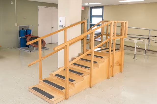 Indoor rehabilitation area with a wooden staircase equipped with handrails and black anti-slip strips on the steps, parallel bars for walking exercises, and exercise equipment against the wall in a well-lit room with beige walls and a tiled floor.