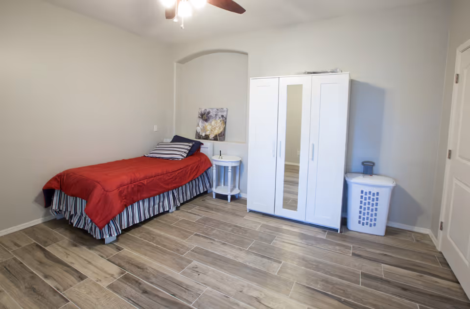 A simple bedroom with a single bed covered with a red blanket and striped bed skirt, a white round side table with a floral artwork resting on it, a white wardrobe with a mirror in the center, and a white laundry basket. The room has wood-look tile flooring and light gray walls.