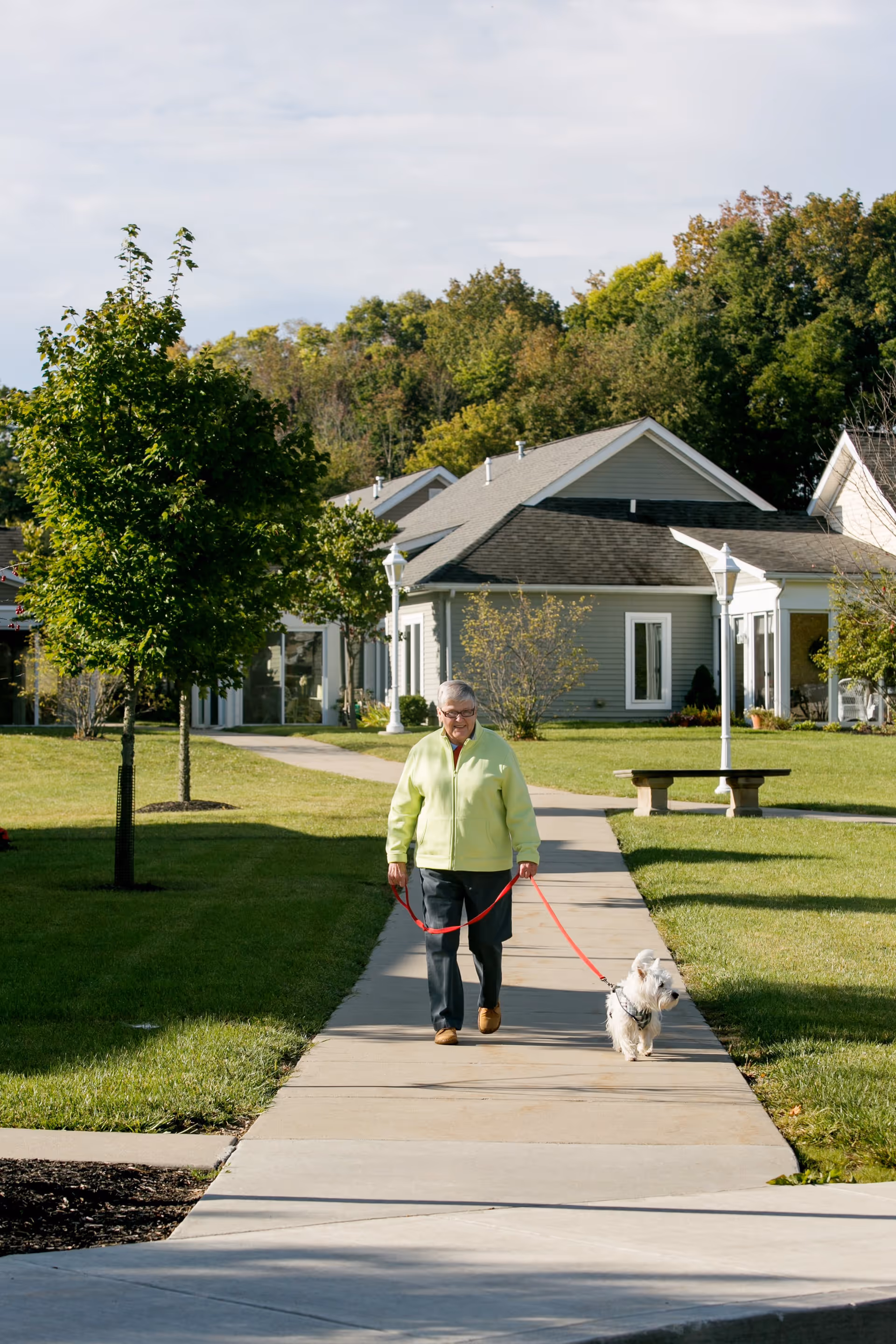 An elderly person wearing a light green jacket walking a small white dog on a red leash along a paved sidewalk in a senior living community with green lawns, trees, and single-story houses in the background.