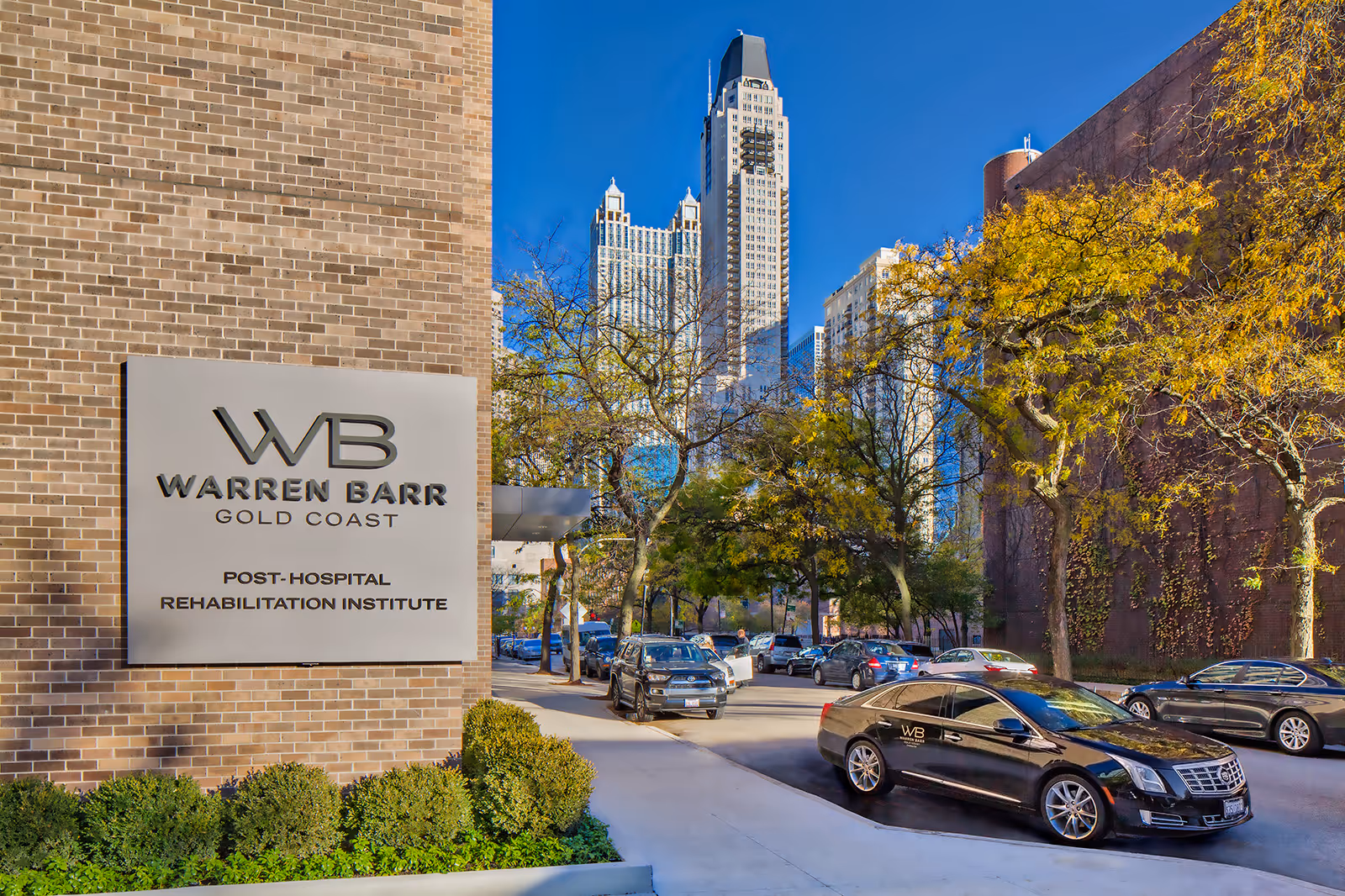 Exterior view of the Warren Barr Gold Coast Post-Hospital Rehabilitation Institute building with a sign on a brick wall. Several cars are parked along the street, including a black car with the WB logo. Trees with autumn foliage line the street and tall city buildings are visible in the background under a clear blue sky.