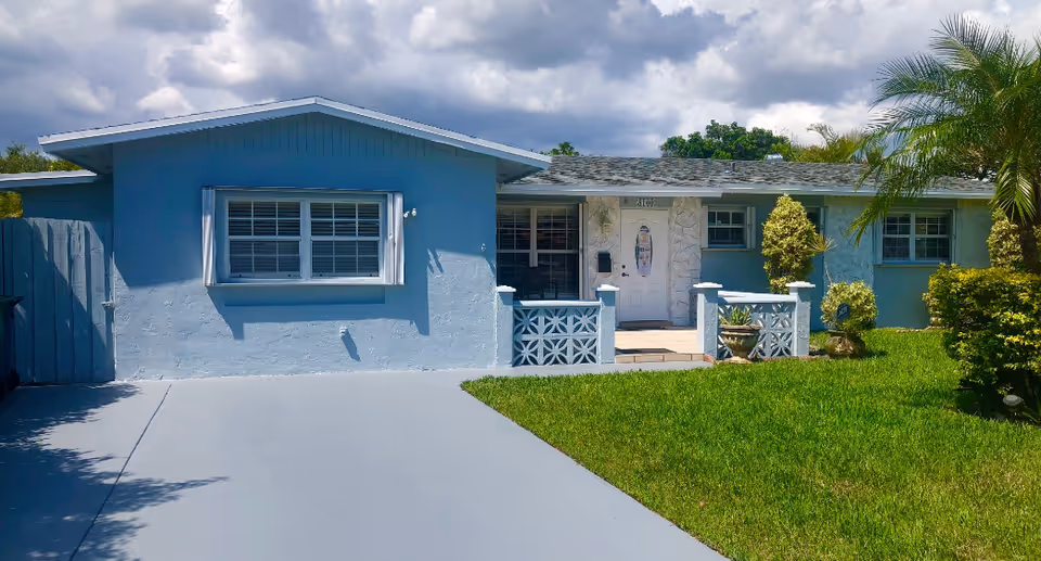 Blue single-story retirement home with a front yard, porch railing, and palm trees.