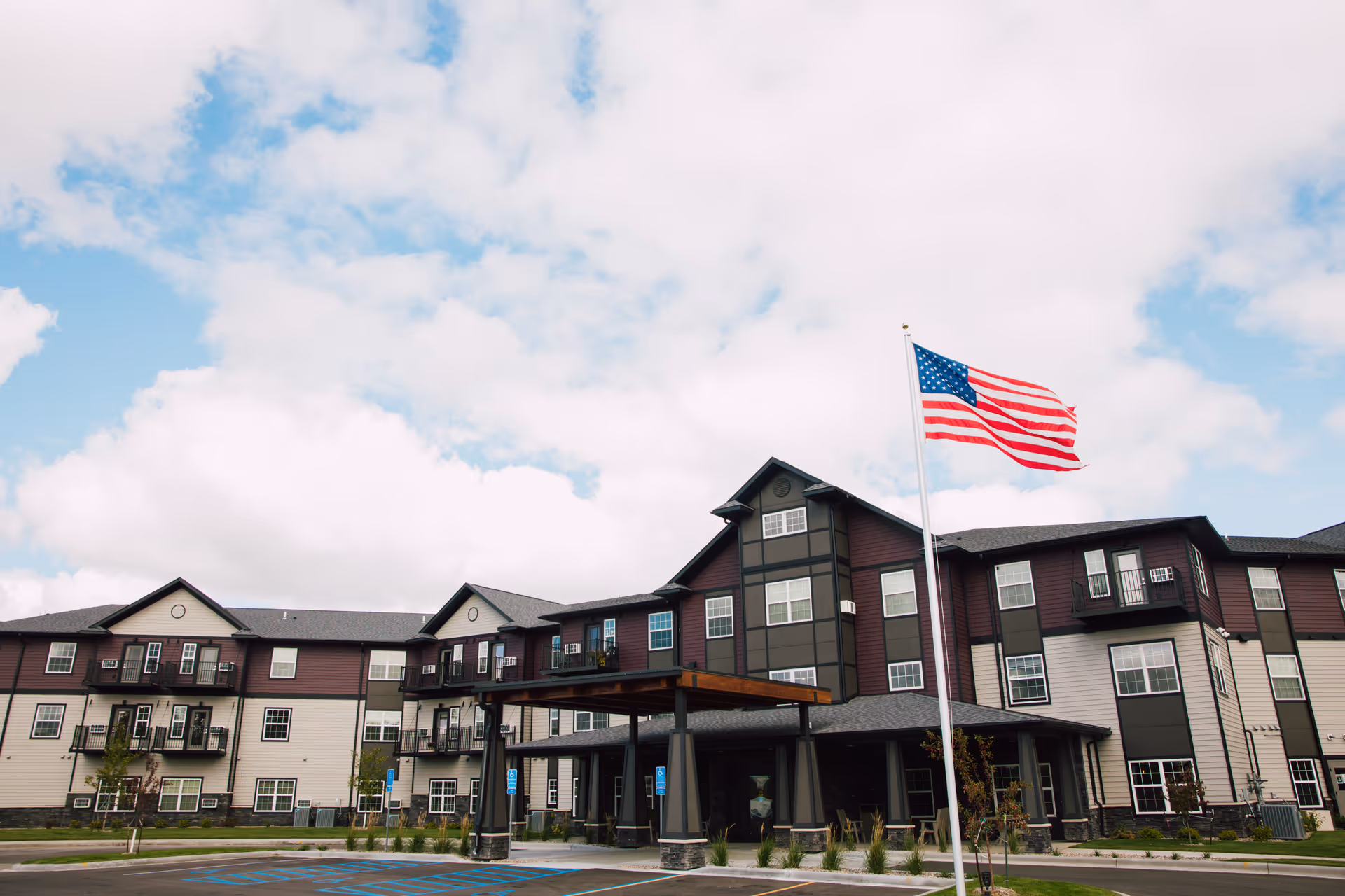 Exterior view of a multi-story senior living facility building with an American flag flying on a flagpole in front. The building has a covered entrance and multiple windows, with a partly cloudy sky in the background.