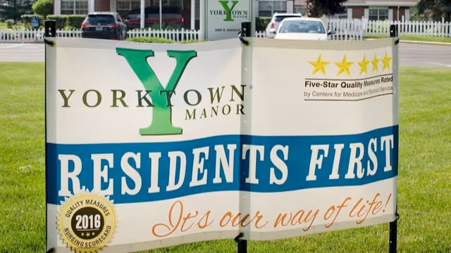 Outdoor sign at Yorktown Manor with the text 'Residents First, It's our way of life!' along with a 2016 Quality Measures Nursing Scorecard badge and a Five-Star Quality Measures Rated by Centers for Medicare & Medicaid Services logo. The sign is placed on a grassy area with parked cars and a white picket fence in the background.
