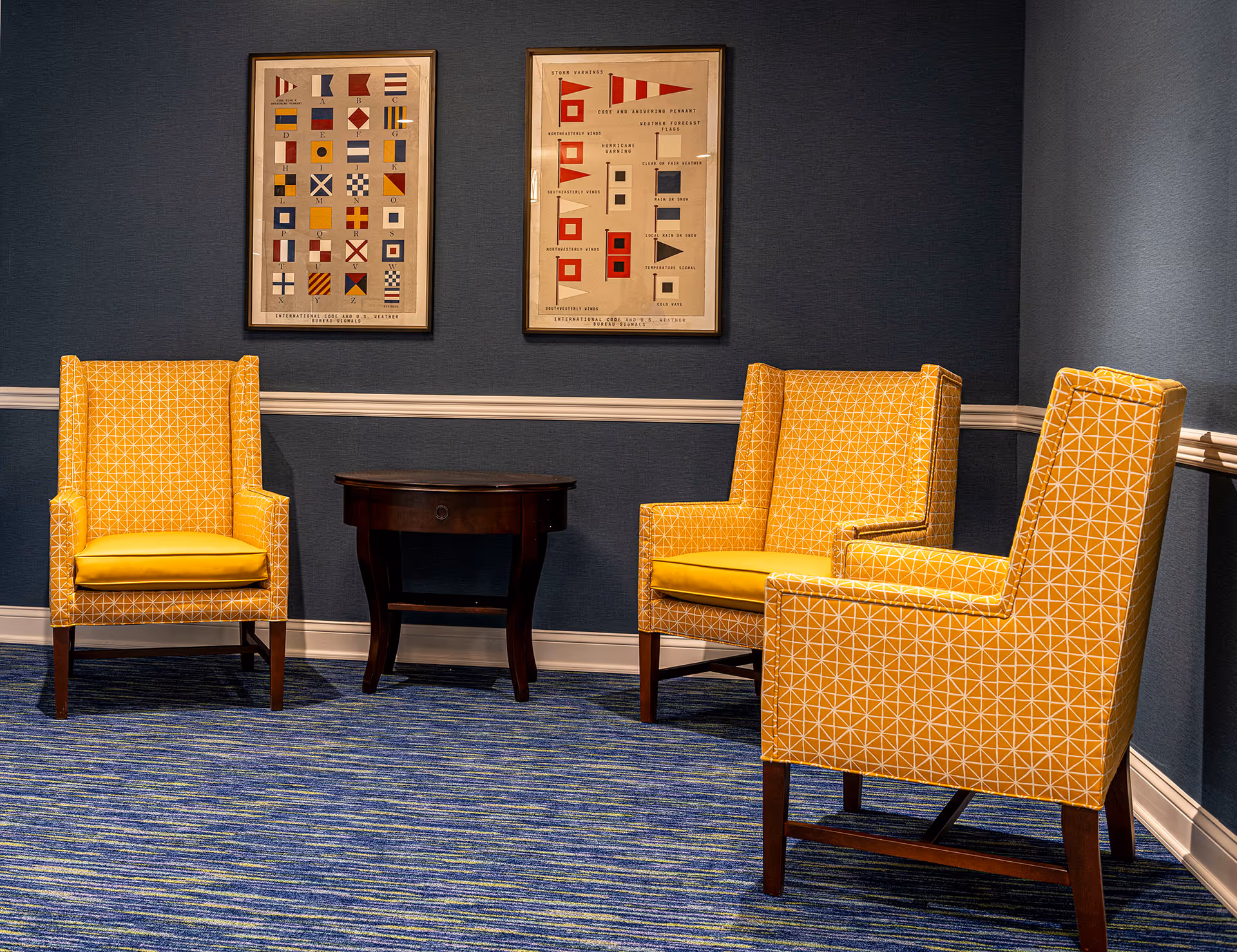A cozy seating area with three yellow patterned armchairs arranged around a small dark wooden side table. The room has blue walls with white trim and two framed nautical signal flag prints hanging on the wall. The floor is covered with a blue carpet featuring green and yellow accents.