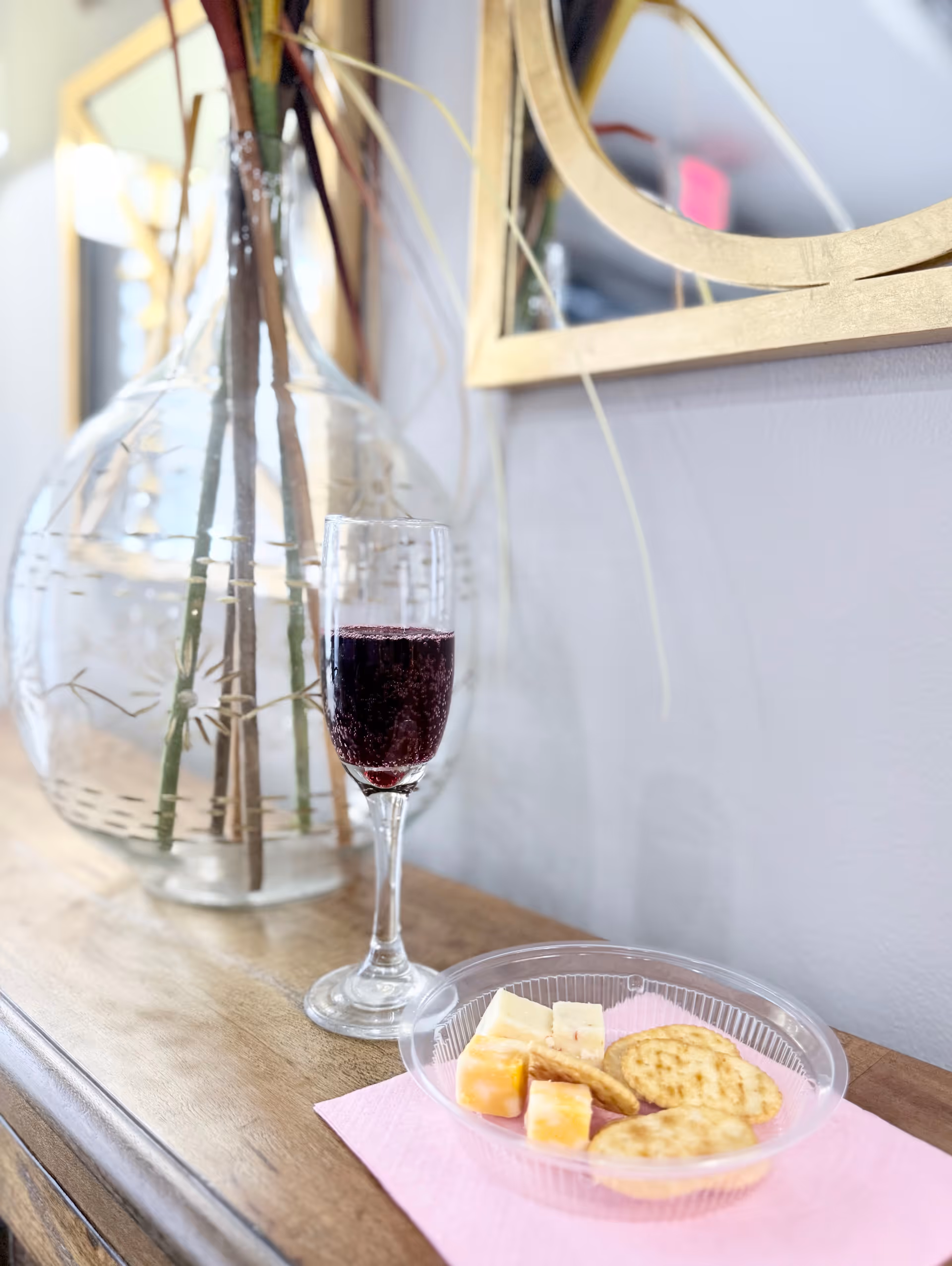 A glass of red wine and a clear plastic plate with assorted cheese cubes and round crackers placed on a wooden surface with a pink napkin. In the background, there is a large clear vase with decorative sticks and a gold-framed mirror on a light-colored wall.