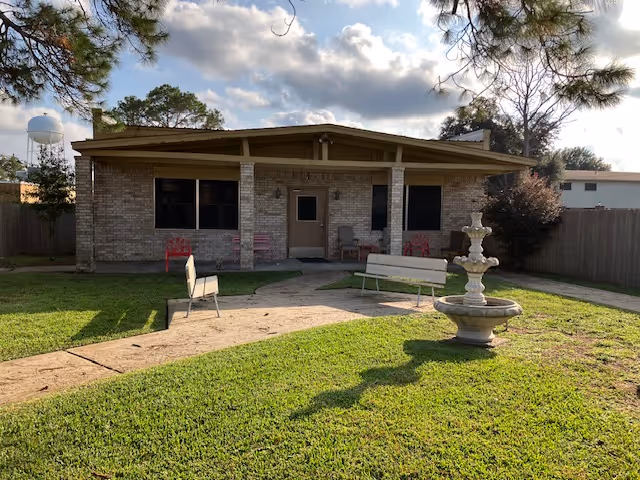 Single-story brick building with a covered porch, benches, a fountain, and a lawn under a partly cloudy sky.