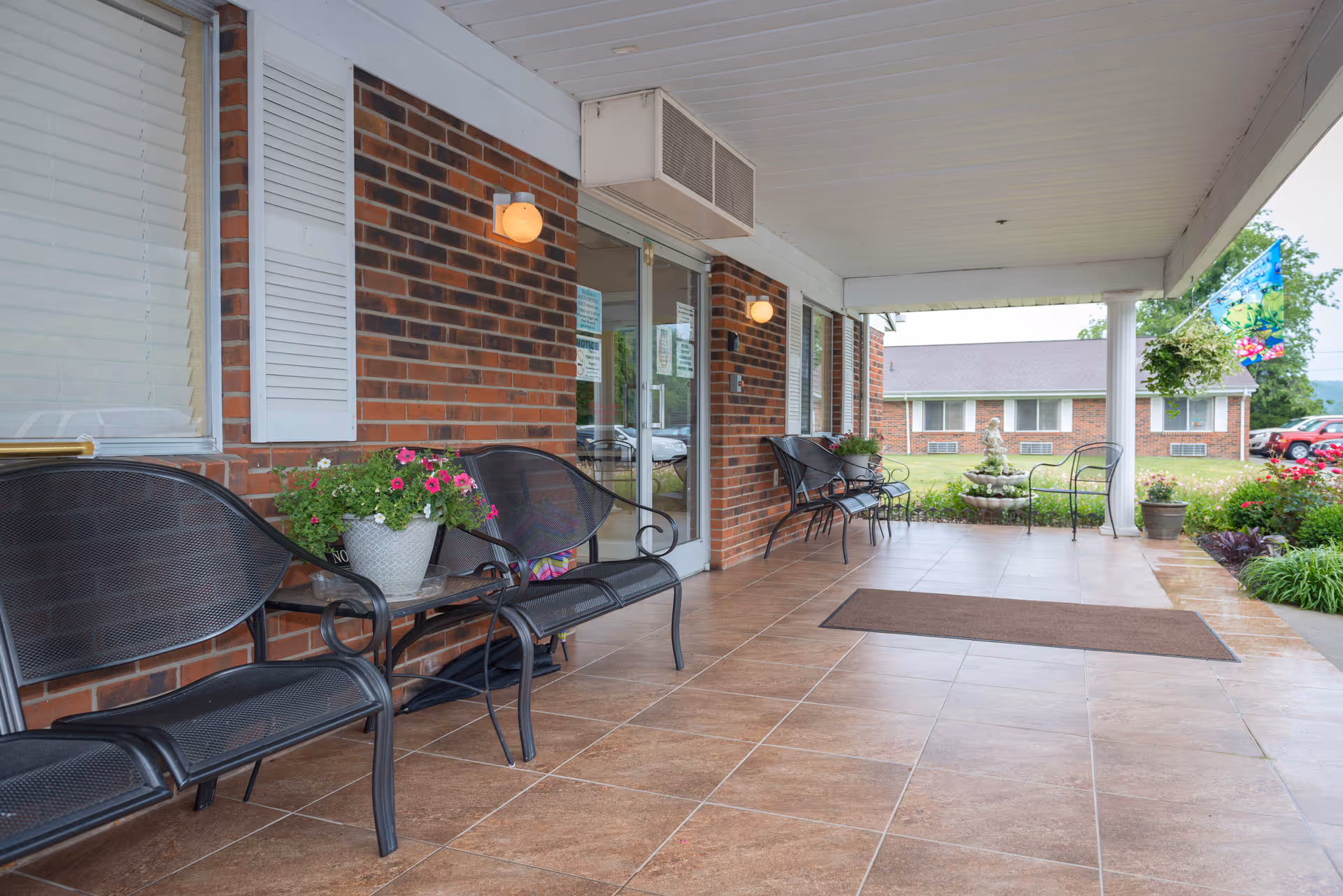Covered outdoor patio area with brown tiled floor, black metal benches, potted flowers, and a brick wall with windows and glass doors. There are two wall-mounted lights and a brown doormat. In the background, there is a grassy area with a small fountain, more potted plants, and a building with multiple windows.